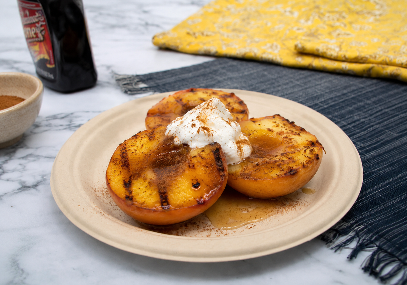 Grilled peaches with whipped cream on a beige plate, placed on a marble surface with a yellow cloth and bottle in the background.